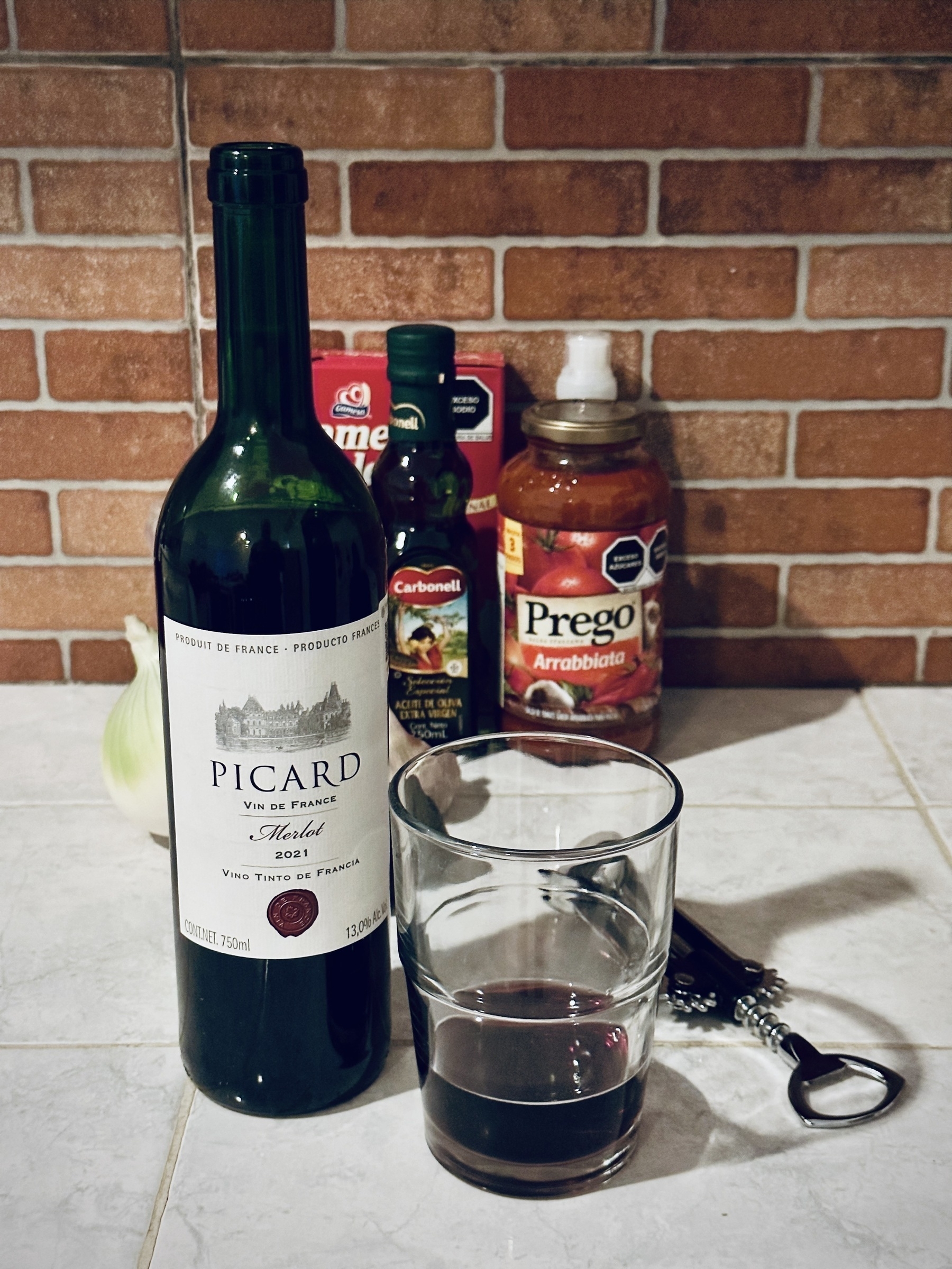 Close up of a quarter full glass tumbler of red wine and the accompanying bottle of Picard Merlot on a white tiled kitchen bench in front of red brown bricks. A bottle opener with ratcheted arms sits just behind. A har of pasta sauce, a small bottle of olive oil, and an onion and a head of garlic line the wall behind.